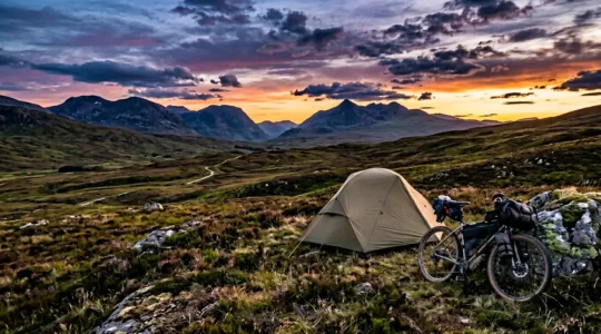 A bikepacker camping beside their loaded bicycle under dramatic twilight sky in remote Scottish wilderness