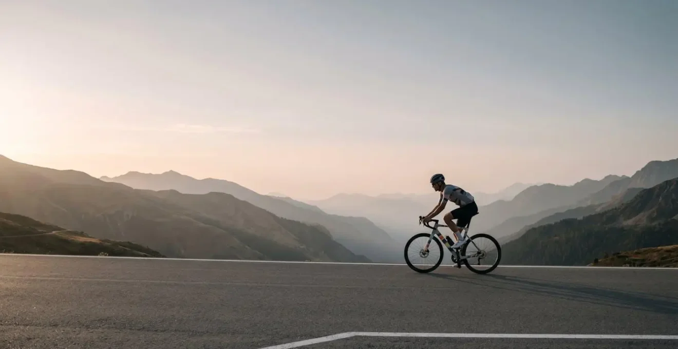 Wide environmental shot of cyclist on winding mountain road with expansive valley view, minimal human presence emphasizing scale and open space